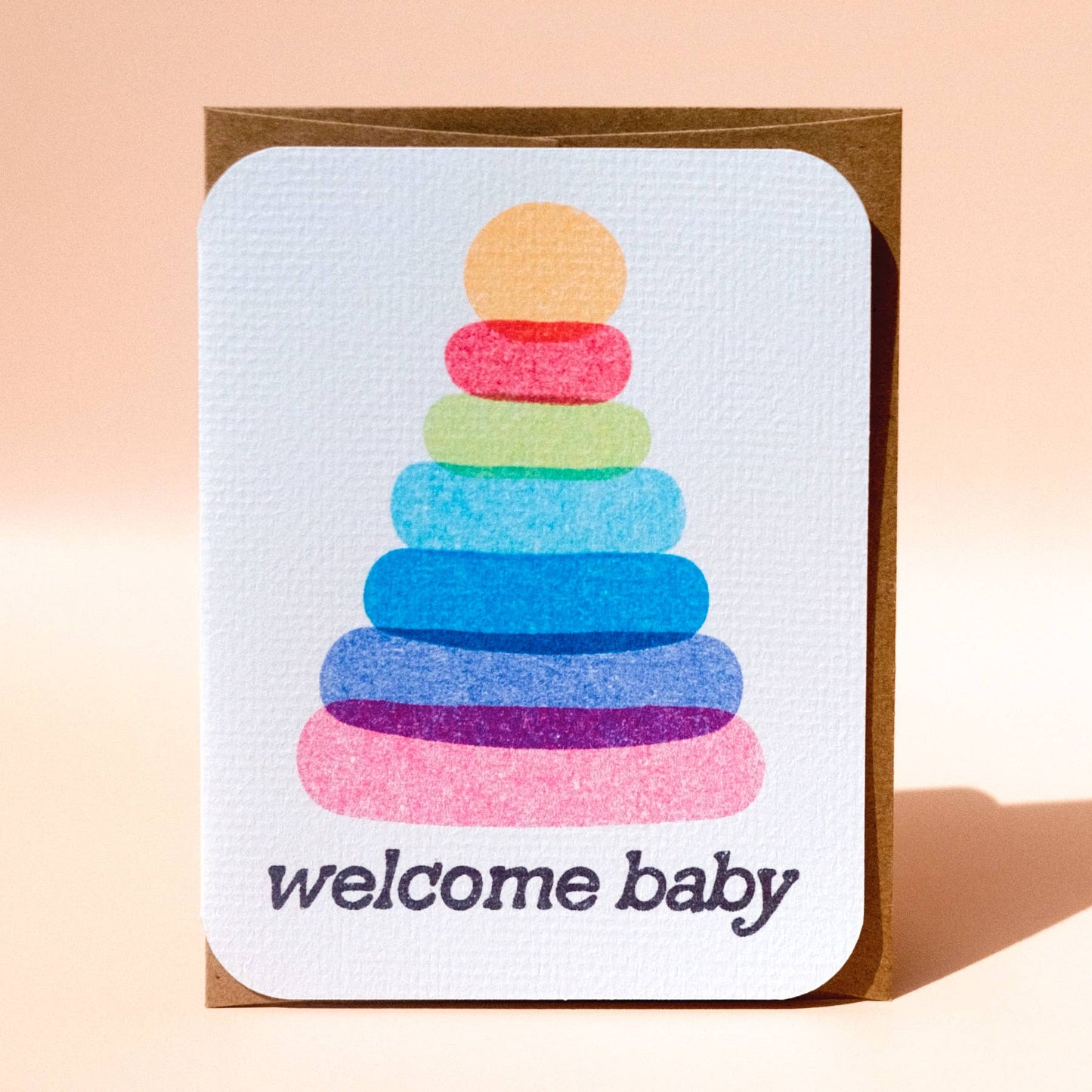 Greeting card with colorful stacking rings design and 'welcome baby' text on a light pink background
