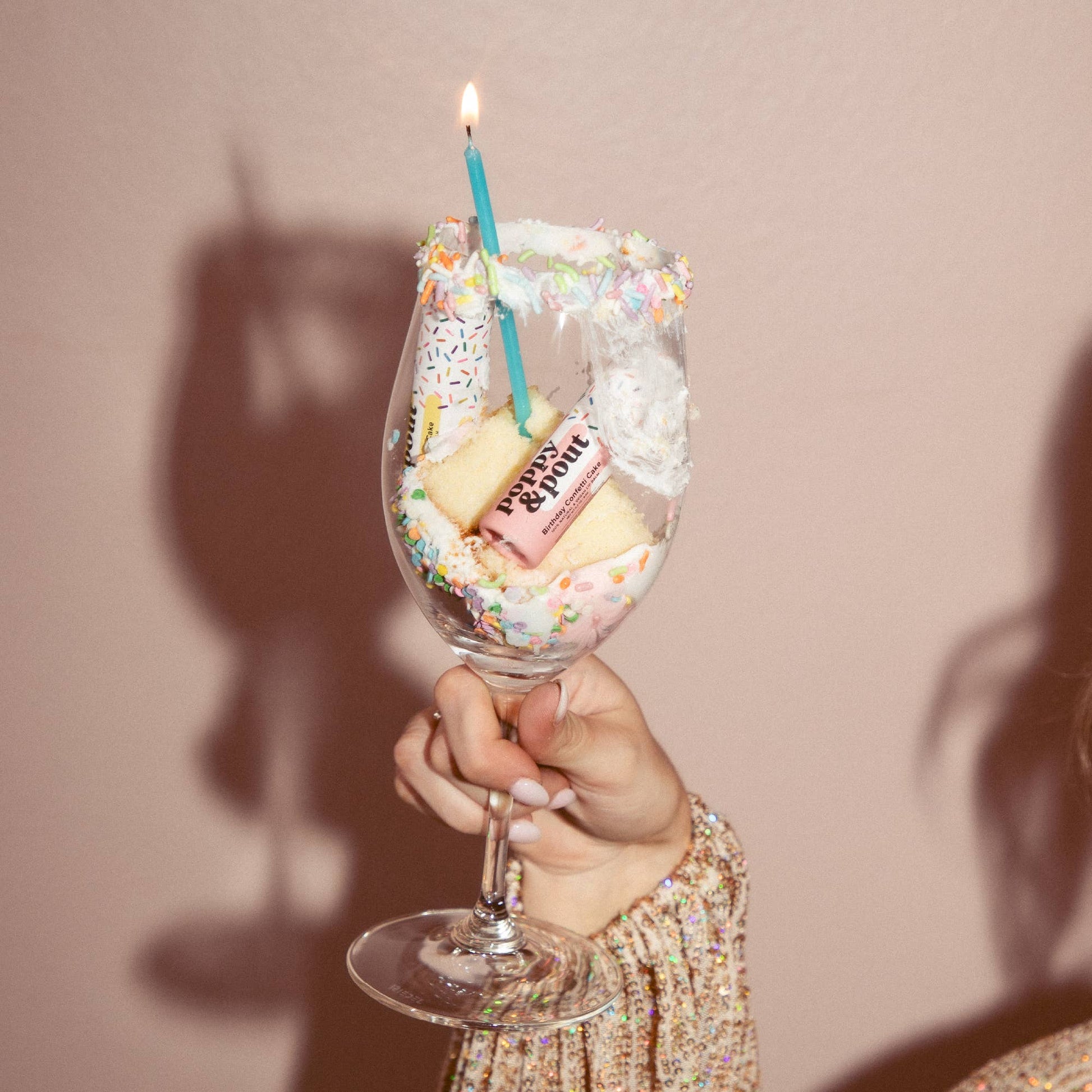 Wine glass filled with cake, candles, and sprinkles held by a person against a plain background
