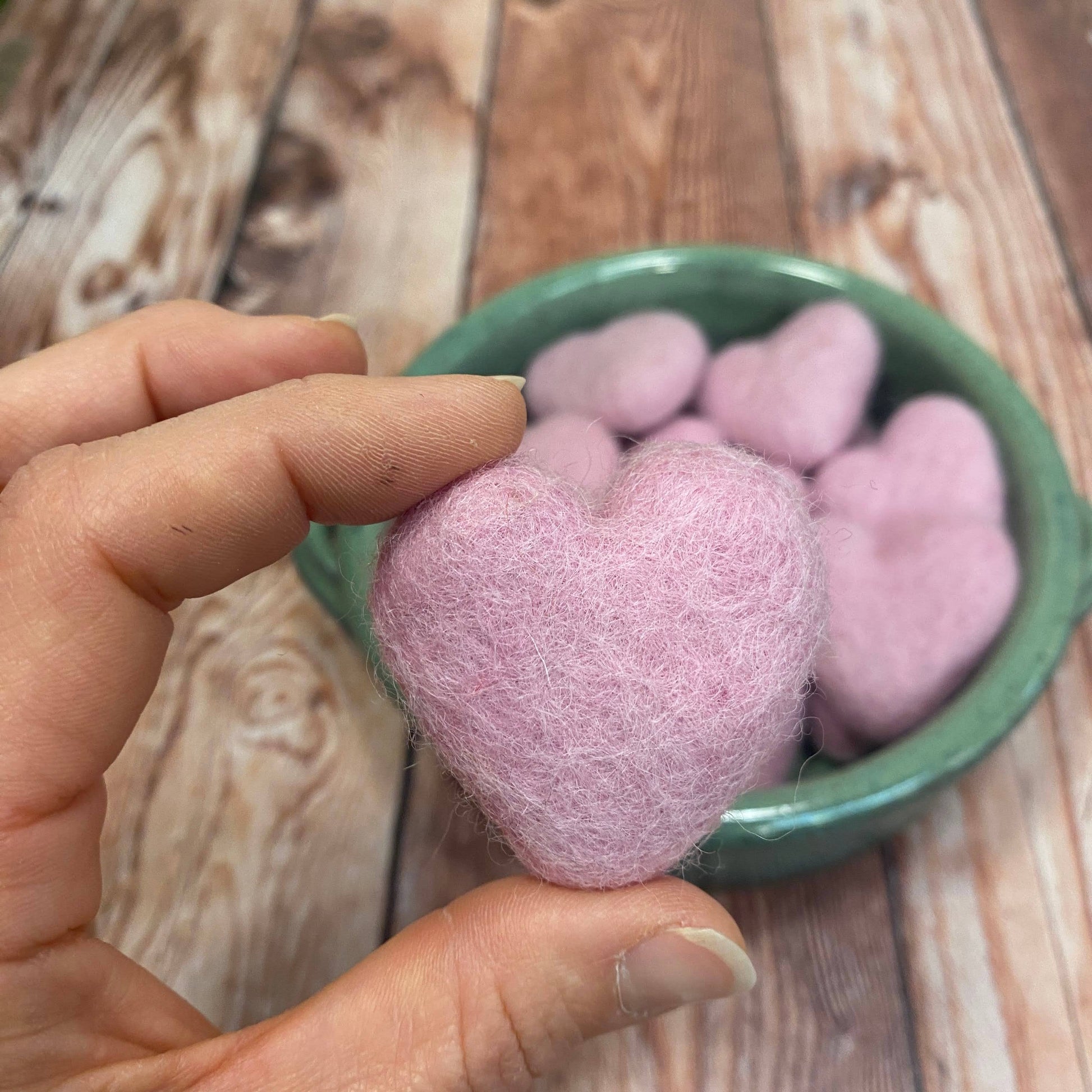 Hand holding a pink heart-shaped object over a green bowl filled with similar objects on a wooden surface.