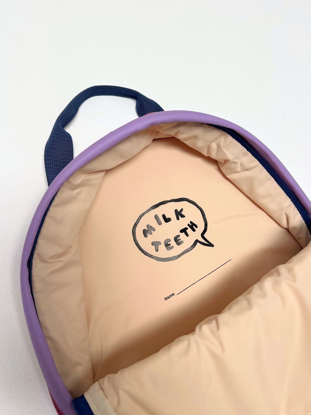 Close-up of the inside of a puffy quilted bag with a light peach interior, purple trim, dark navy handles, and a printed speech-bubble logo reading 'MILK TEETH' on the lining.