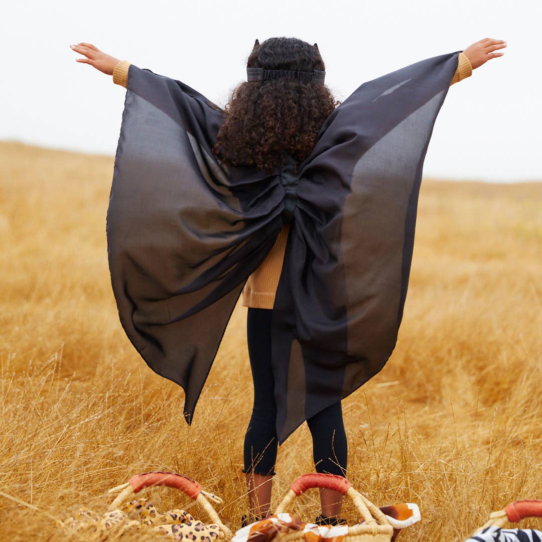 A woman stands with her back to the camera in a golden field, wearing a sheer black cape-like wrap draped over her shoulders and held out wide with her arms. Two woven baskets with orange handles sit on the ground in front of her.