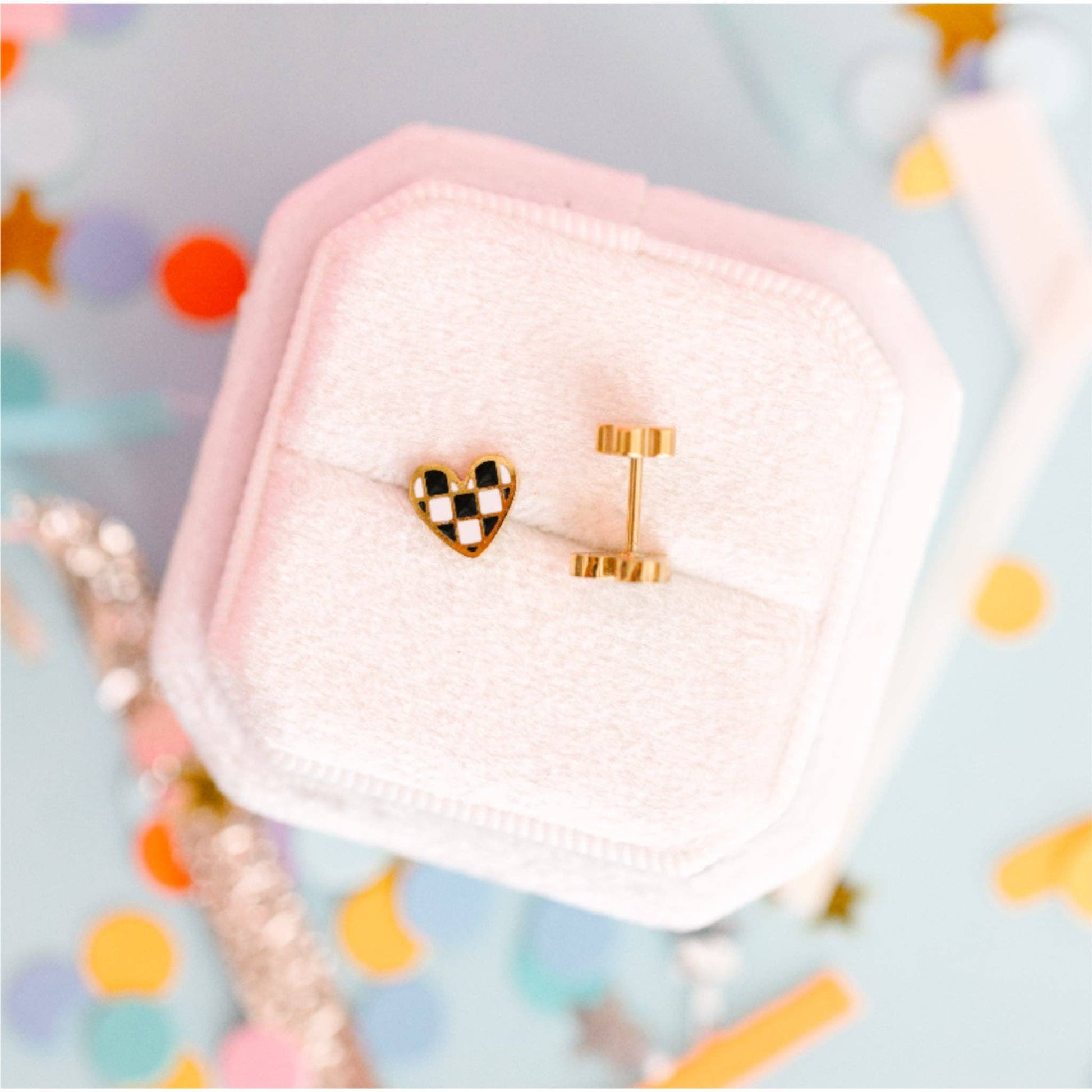 Gold earrings with a checkered heart design in a pink jewelry box on a colorful confetti background
