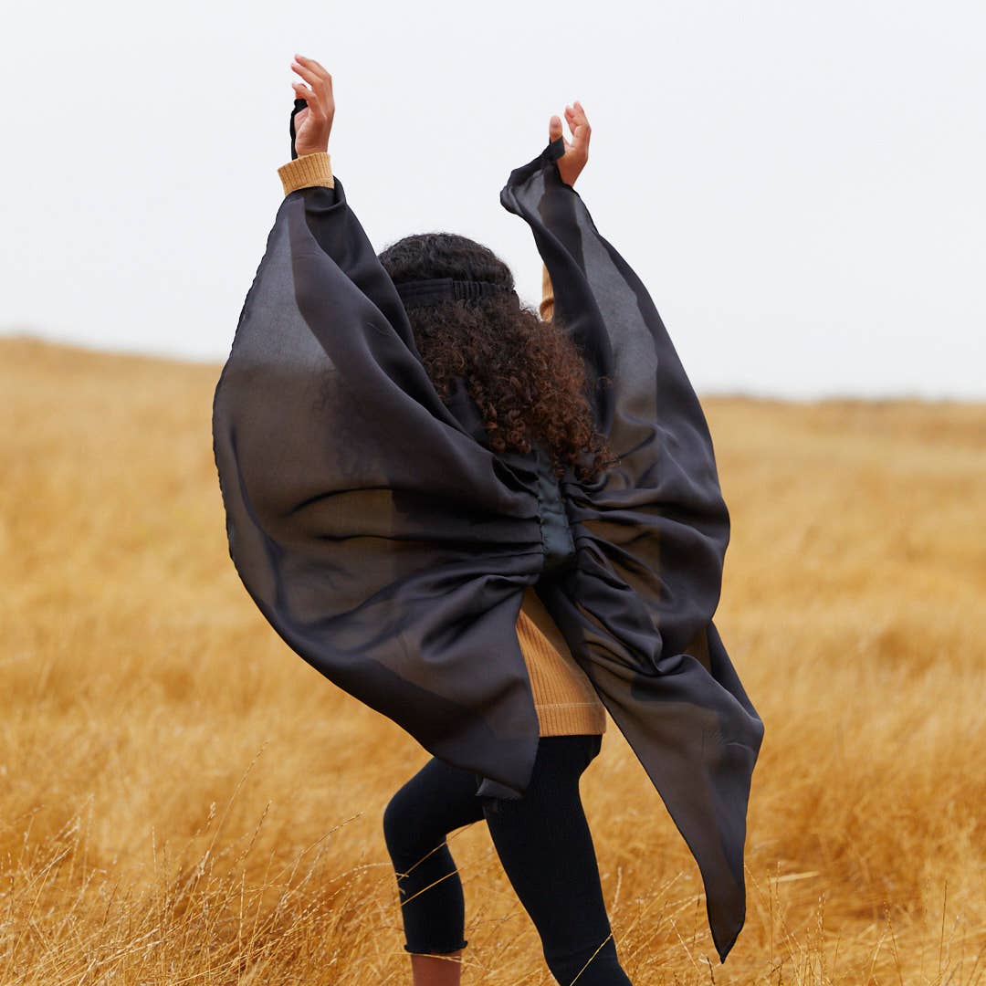 A person standing in a golden field with their back to the camera, wearing a large flowing black bat wings 