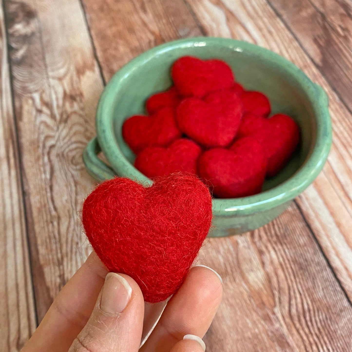 Red heart-shaped object held by a hand with a green bowl filled with similar hearts on a wooden surface.
