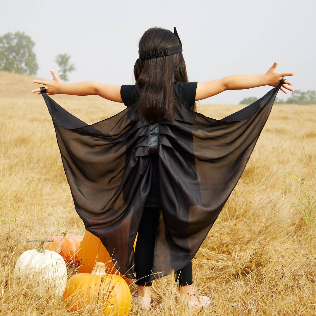 A child standing in a dry grass field with arms outstretched, wearing a black bat cape and a black headband, with pumpkins at the bottom left.